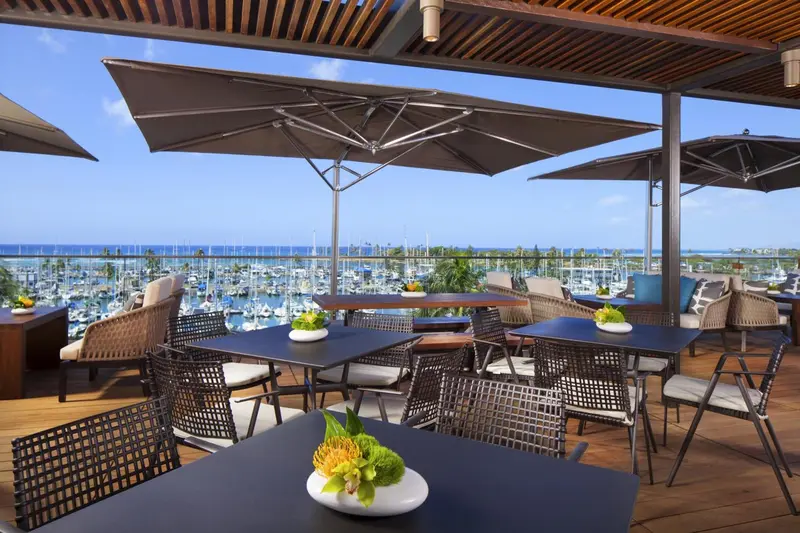 Terrasse ombragée avec tables et chaises donnant sur un port de plaisance.