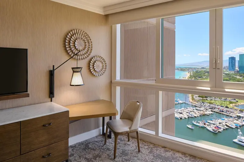 Une chambre lumineuse avec un bureau près d'une grande fenêtre offrant une vue sur la mer et un port de plaisance.