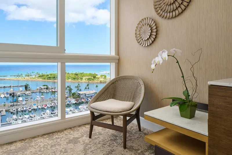 Un fauteuil en osier est placé devant une grande fenêtre donnant sur la mer et un port de plaisance.