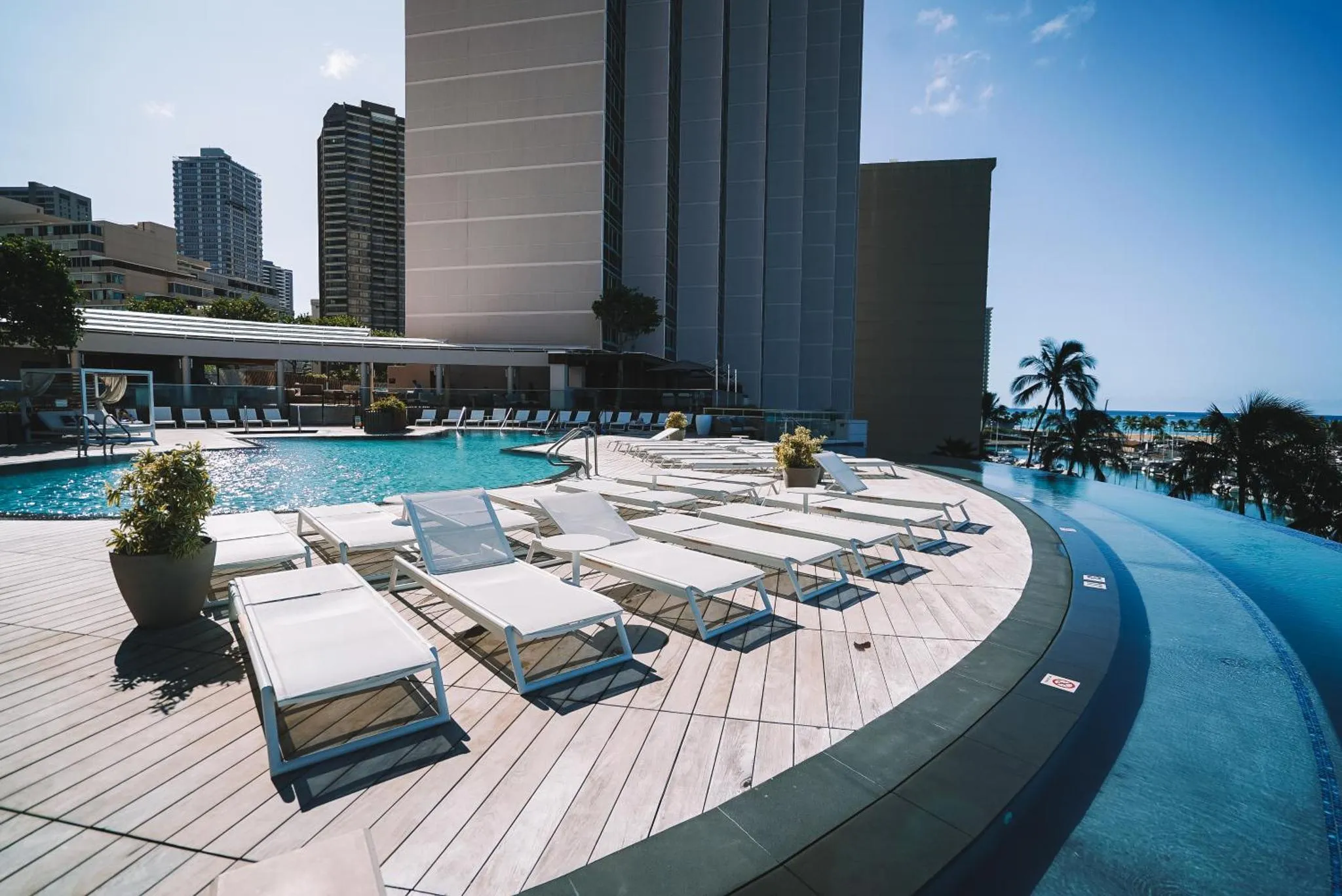 Transats blancs disposés autour d'une piscine à débordement avec vue sur la mer et la ville.