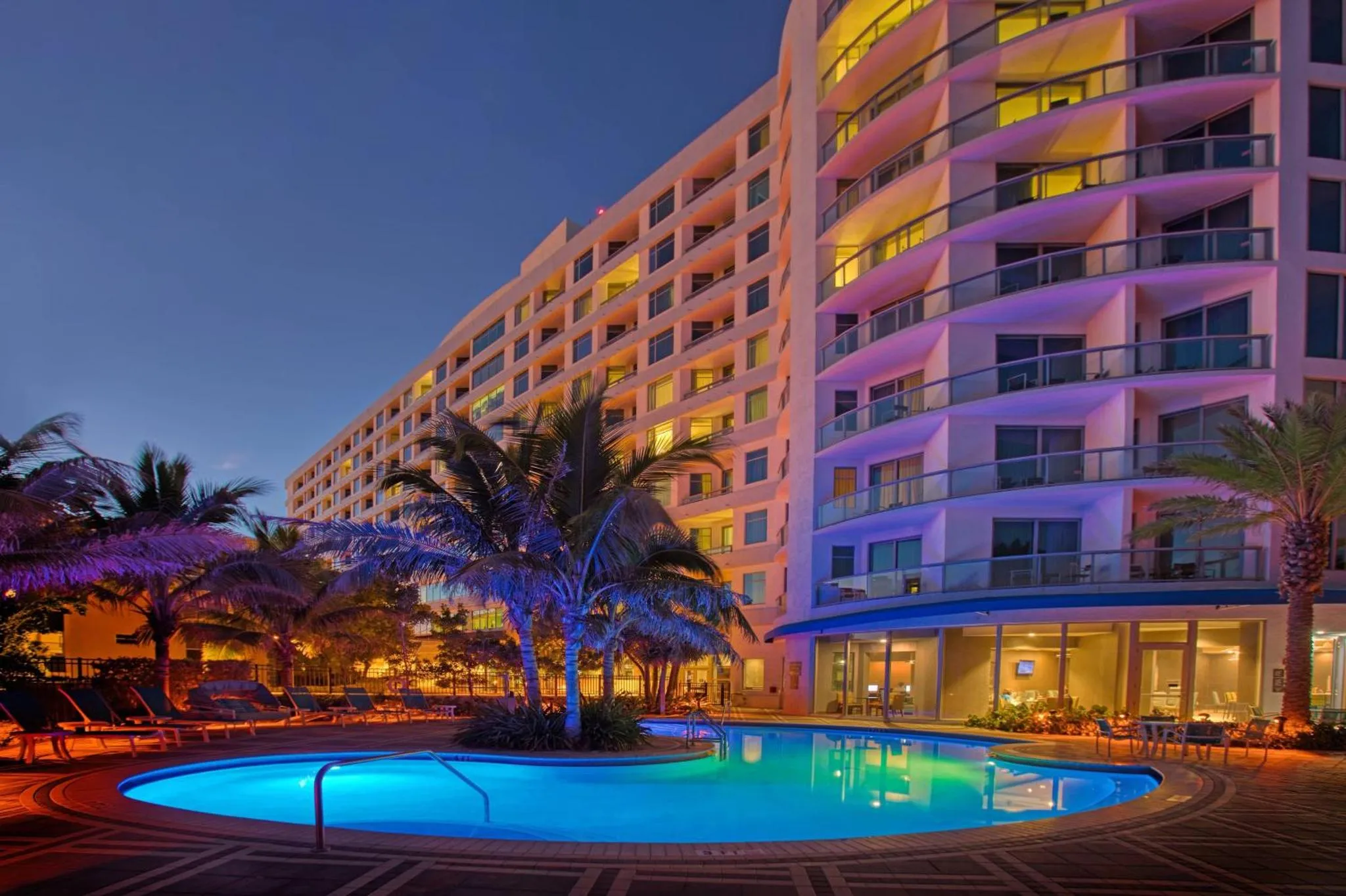Bâtiment illuminé avec piscine au premier plan et palmiers.