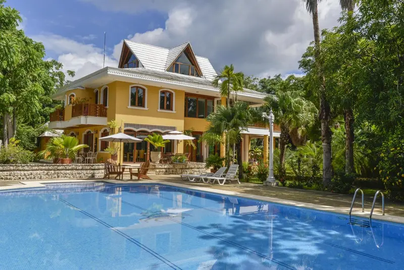 Une grande villa jaune et blanche avec une piscine au premier plan et une végétation luxuriante.