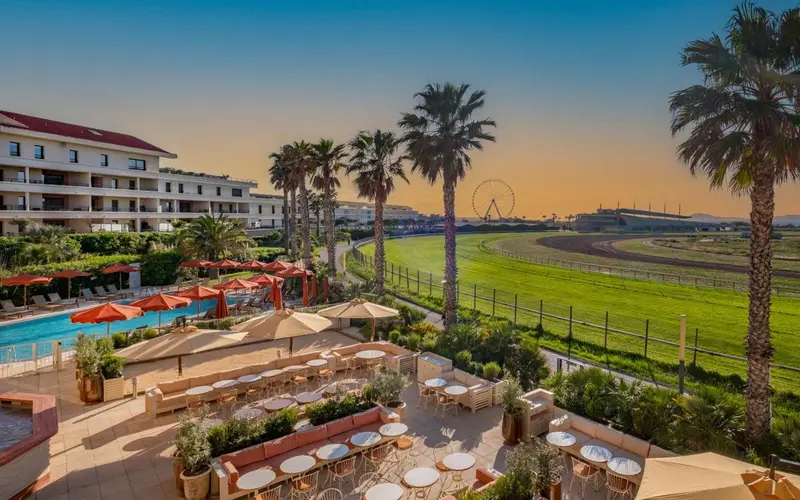 Terrasse ombragée avec tables et chaises, devant un bâtiment et une piscine.