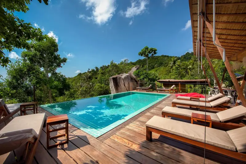 Transats et piscine à débordement avec végétation luxuriante en arrière-plan.