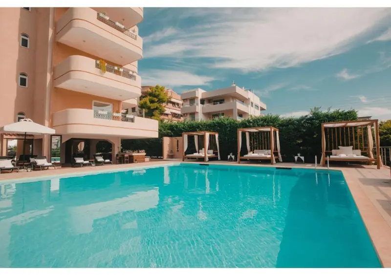 Une piscine turquoise bordée de transats blancs et de parasols, devant un bâtiment beige.