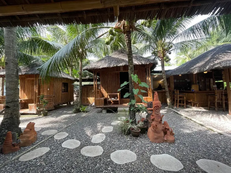 Des bungalows en bois sont alignés dans une cour gravillonnée ombragée par des palmiers.