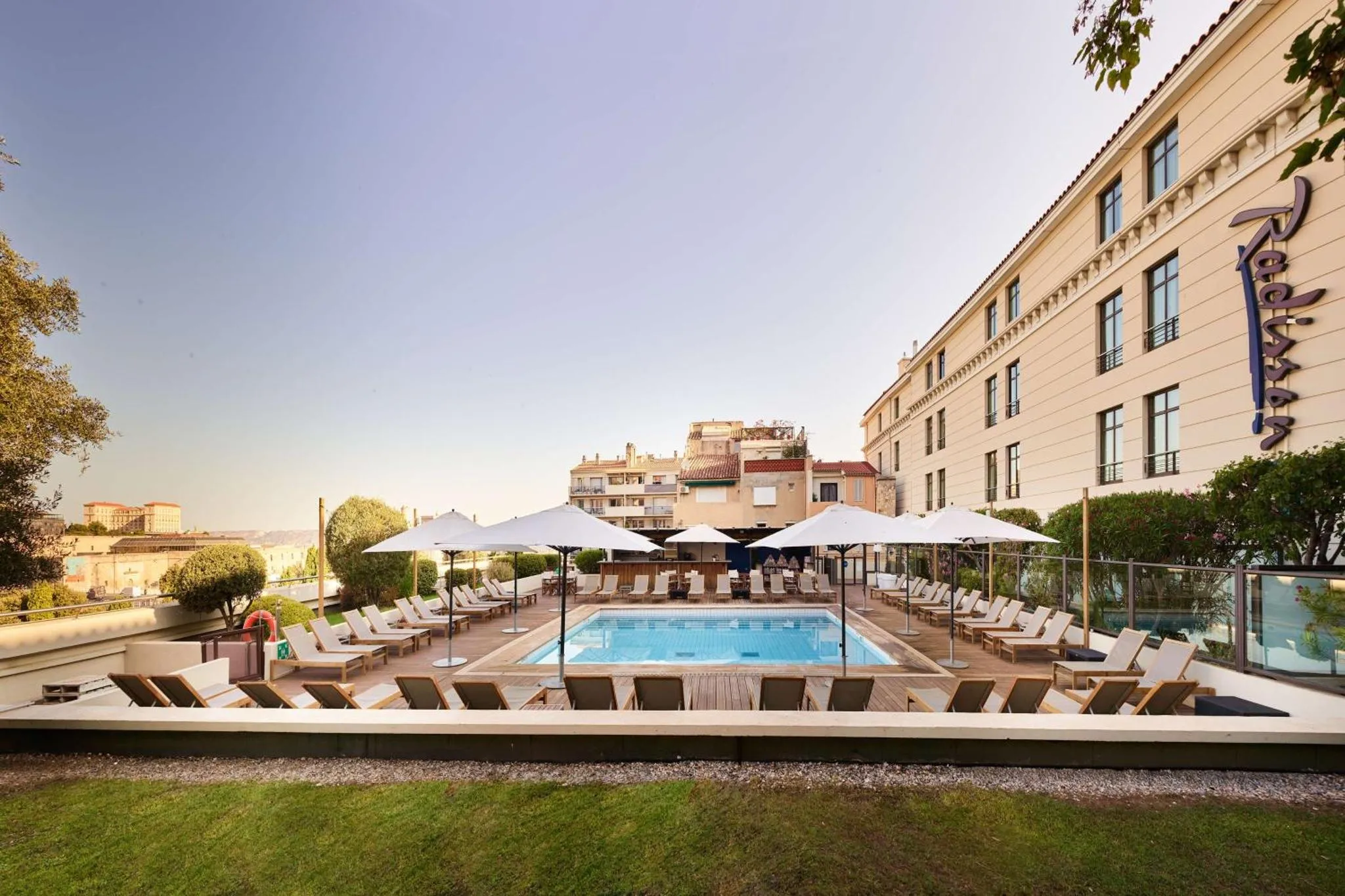 Une piscine rectangulaire entourée de transats et de parasols, devant un bâtiment beige.