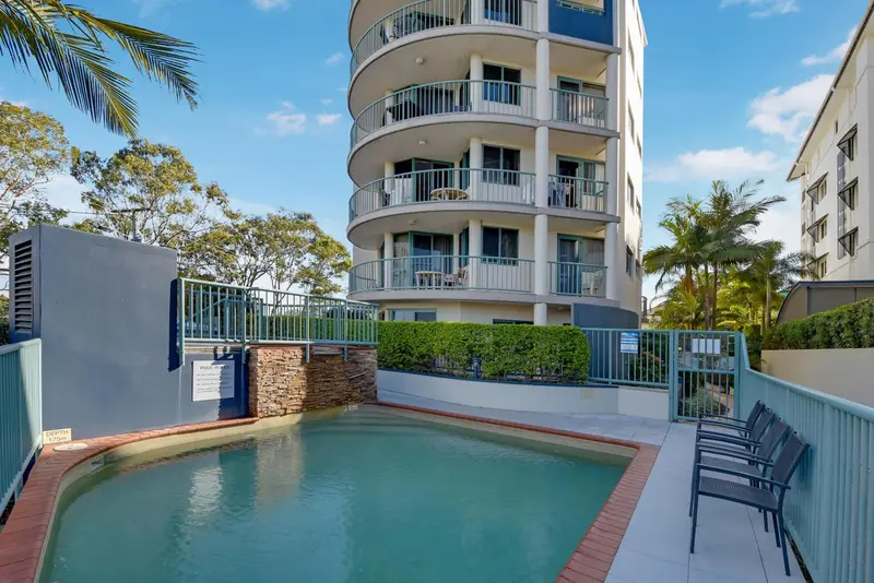 Une piscine turquoise au premier plan, devant un bâtiment résidentiel avec des balcons.