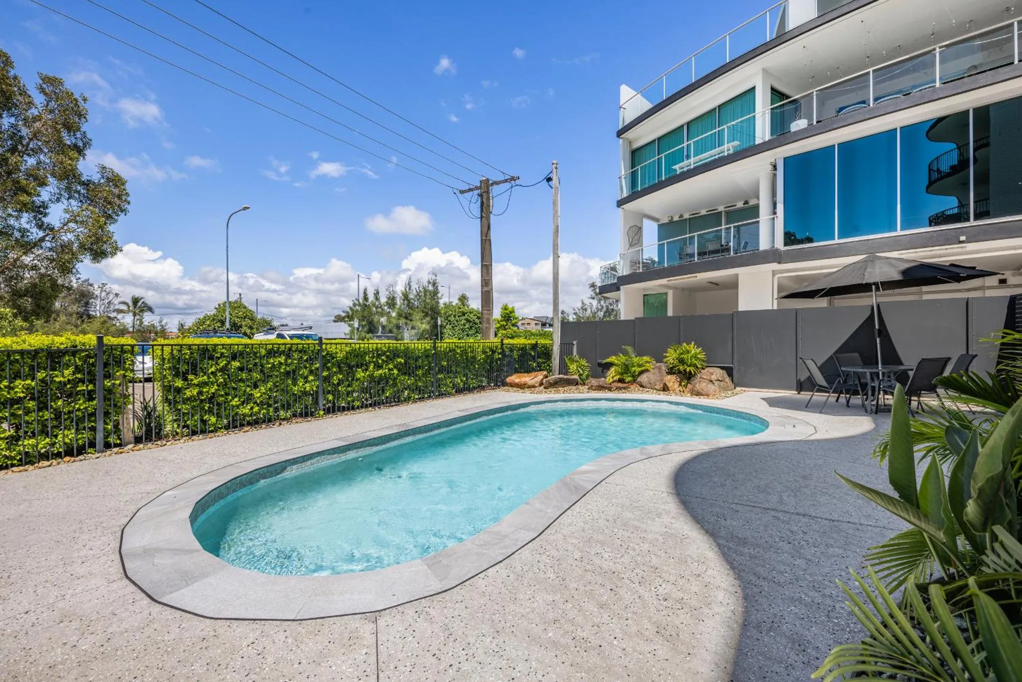 Une piscine rectangulaire bordée d'une terrasse ensoleillée devant un bâtiment moderne.