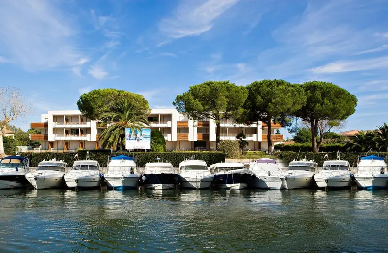 Bâtiment blanc bordé de pins et de bateaux amarrés dans un port.