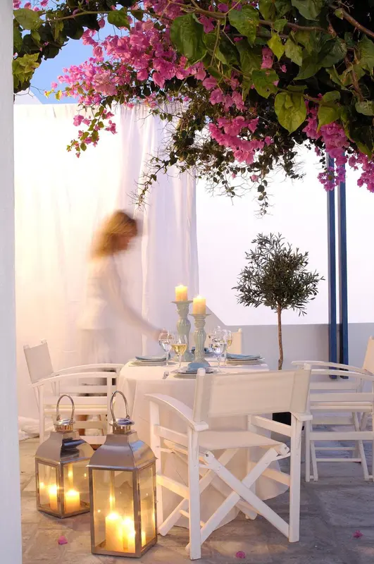 Une table blanche est dressée sur une terrasse ombragée par des fleurs roses.