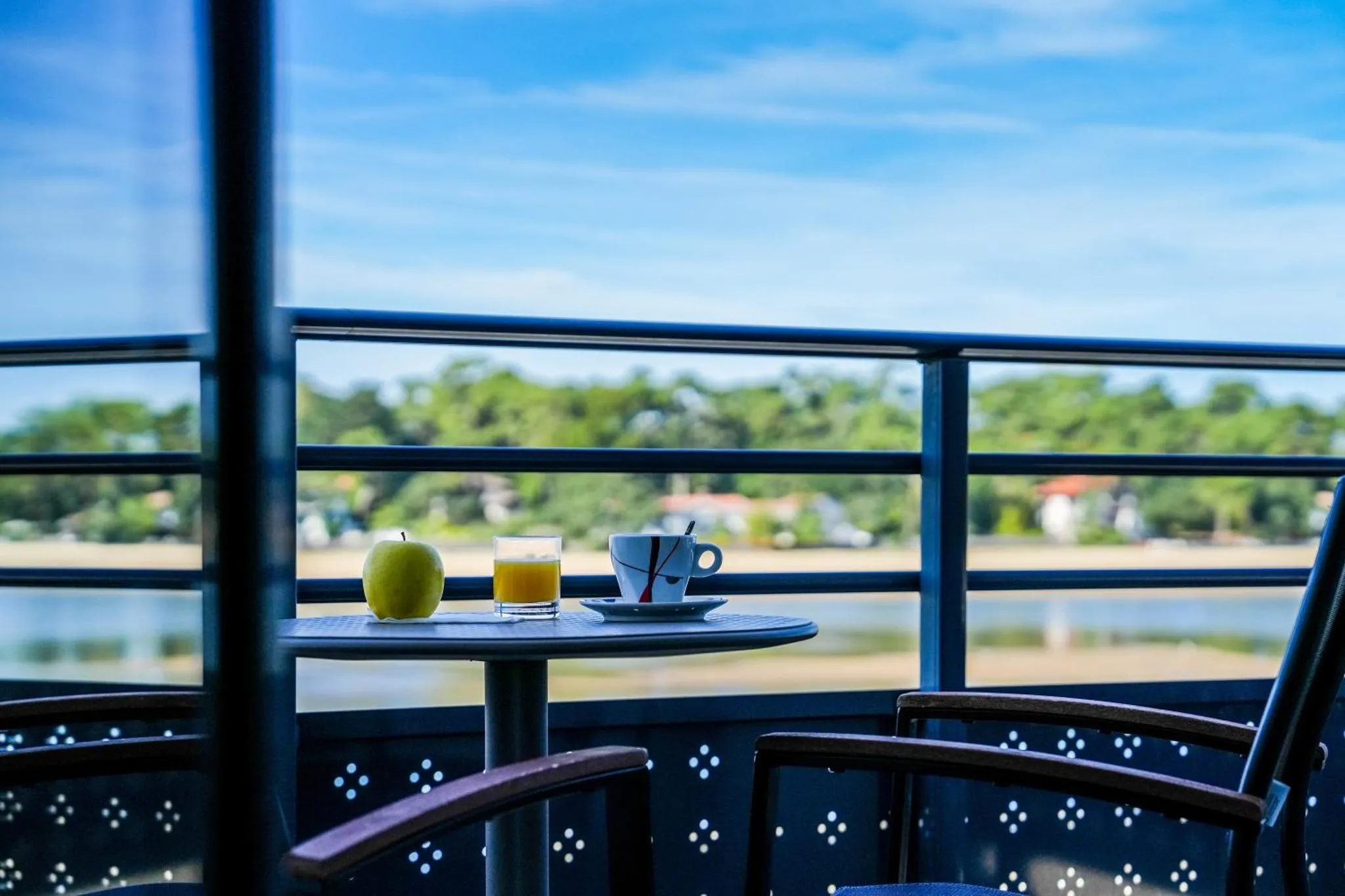 Une table avec des boissons et des fruits est posée sur un balcon avec vue sur la mer et la végétation.