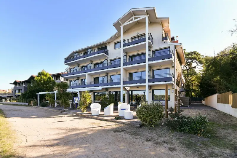 Bâtiment blanc à plusieurs étages avec des balcons et une végétation environnante.