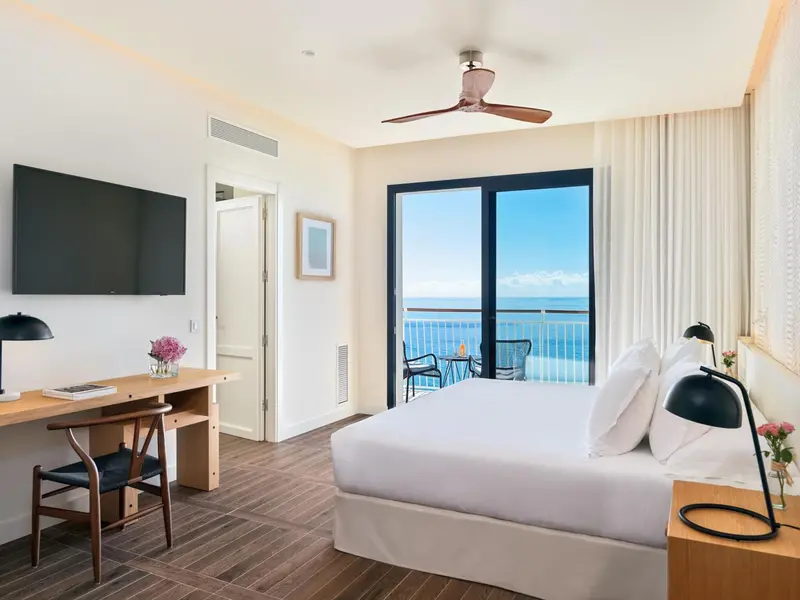 Chambre lumineuse avec un lit blanc et un bureau près d'une baie vitrée donnant sur la mer.