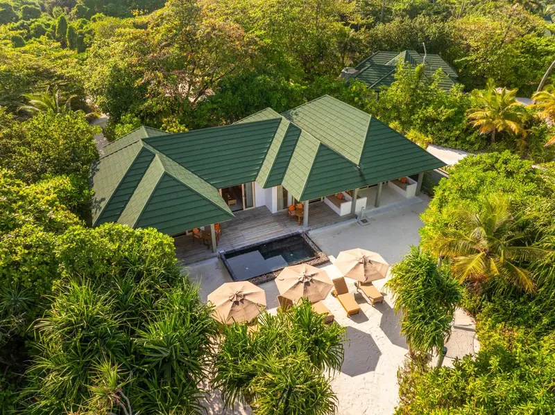 Vue aérienne d'un complexe hôtelier tropical avec des villas aux toits verts et des transats sur une plage de sable blanc.