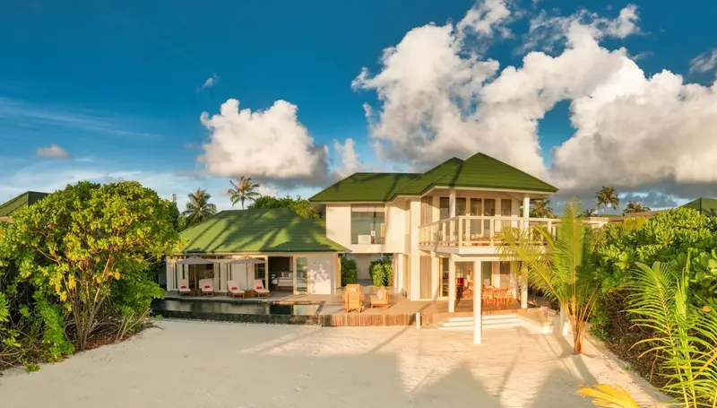 Villa blanche avec toiture verte et terrasse ensoleillée face à une plage de sable fin.