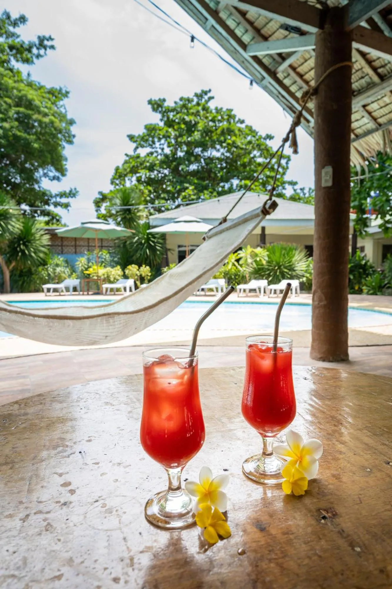 Deux cocktails colorés reposent sur une table en bois près d'une piscine et d'un hamac.