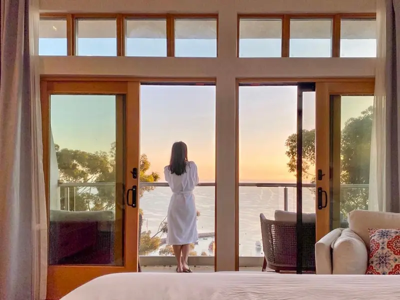 Une personne se tient devant une baie vitrée ouverte sur une vue maritime au lever du soleil.