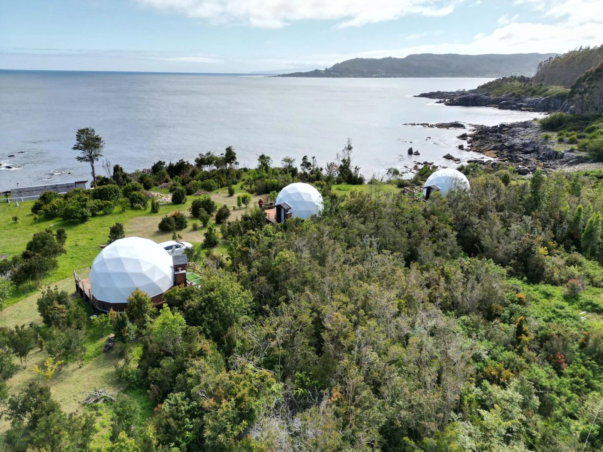 Vue aérienne de structures géodésiques blanches sur une colline verdoyante surplombant la mer.