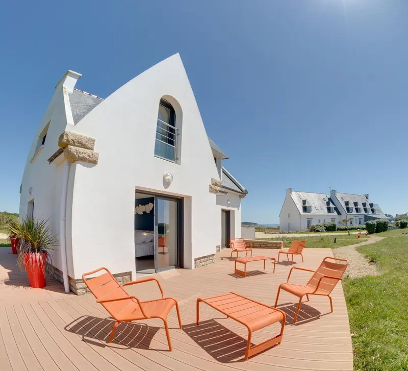Façade blanche d'une maison au toit pointu avec une terrasse en bois et des chaises longues orange.