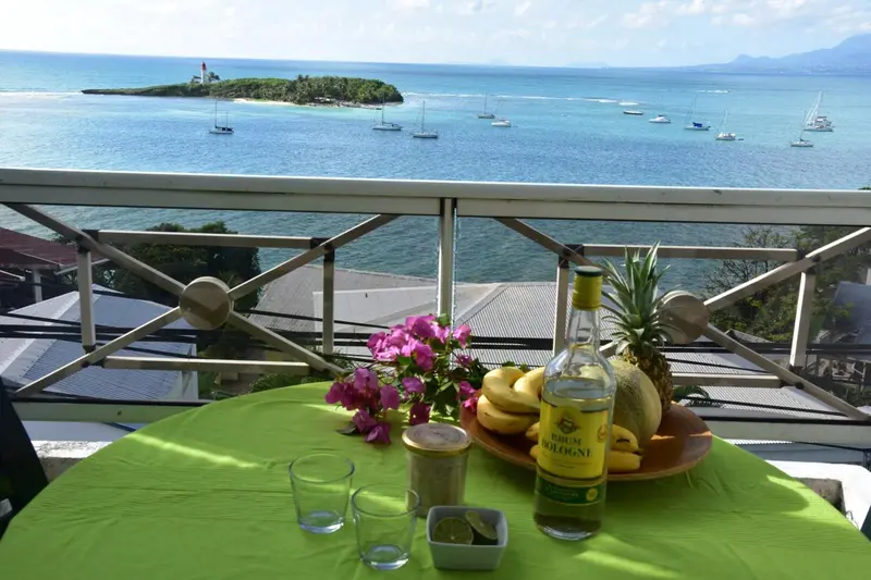 Une table verte avec une bouteille, des fruits et des verres est mise en scène sur un balcon avec vue sur la mer.