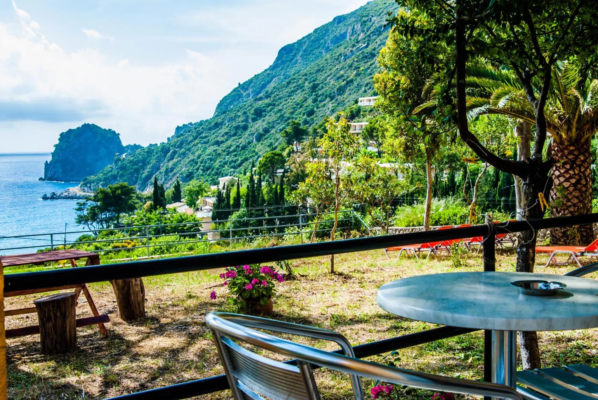 Tables et chaises sur une terrasse ensoleillée avec vue sur la mer et les montagnes.