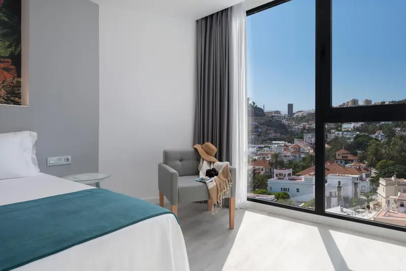Une chambre lumineuse avec un lit et un fauteuil près d'une grande fenêtre offrant une vue sur la ville.