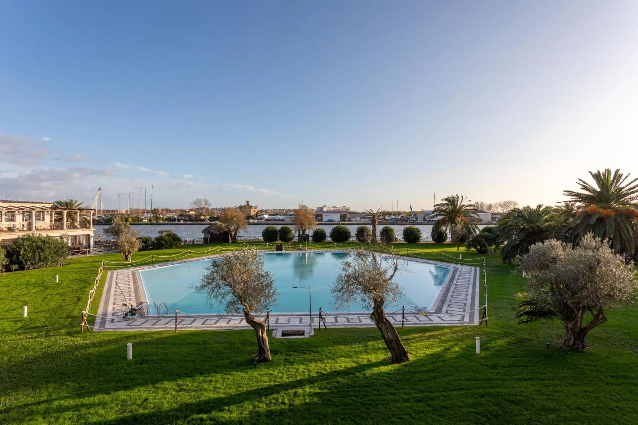 Une piscine rectangulaire bordée d'herbe verdoyante, avec des arbres et une vue sur un port en arrière-plan.