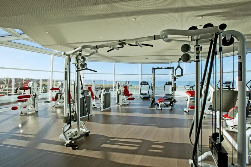 Une salle de sport lumineuse avec des équipements de musculation face à une grande baie vitrée donnant sur la mer.