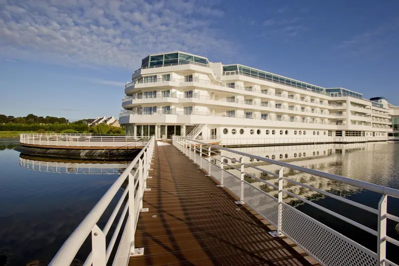 Bâtiment blanc moderne avec des balcons surplombant l'eau calme et un ponton en bois.