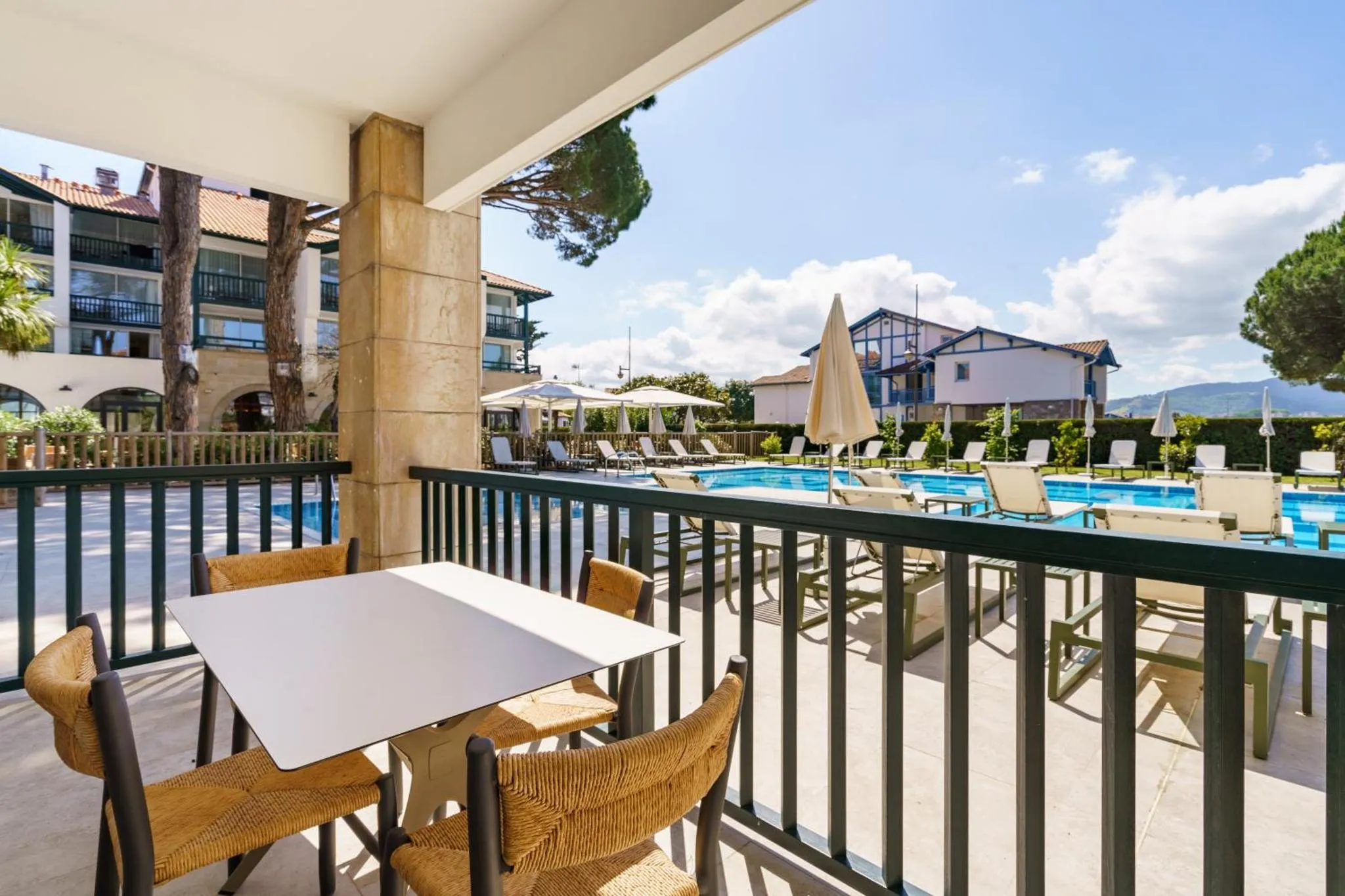 Une terrasse ombragée avec tables et chaises donne sur une piscine et un jardin.