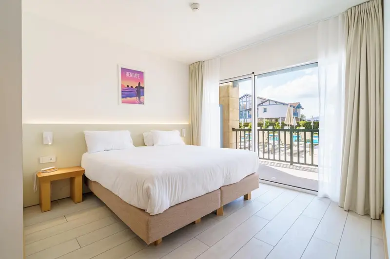 Chambre lumineuse avec un grand lit et vue sur une piscine.