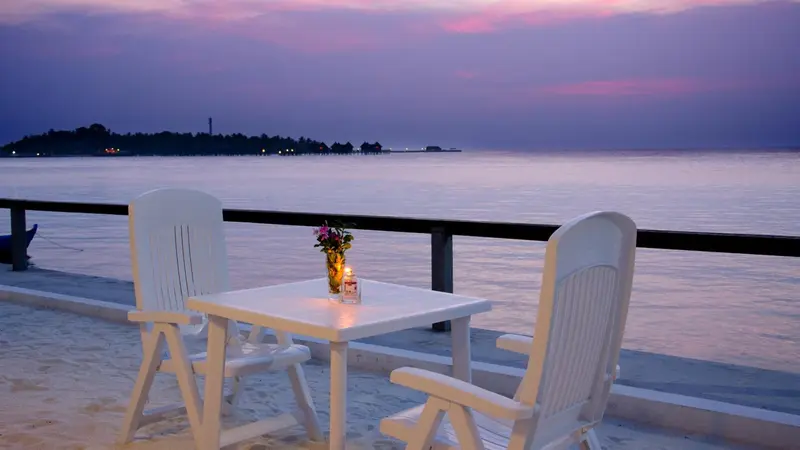 Une table et deux chaises en plastique blanc sont disposées sur une terrasse face à la mer au coucher du soleil.