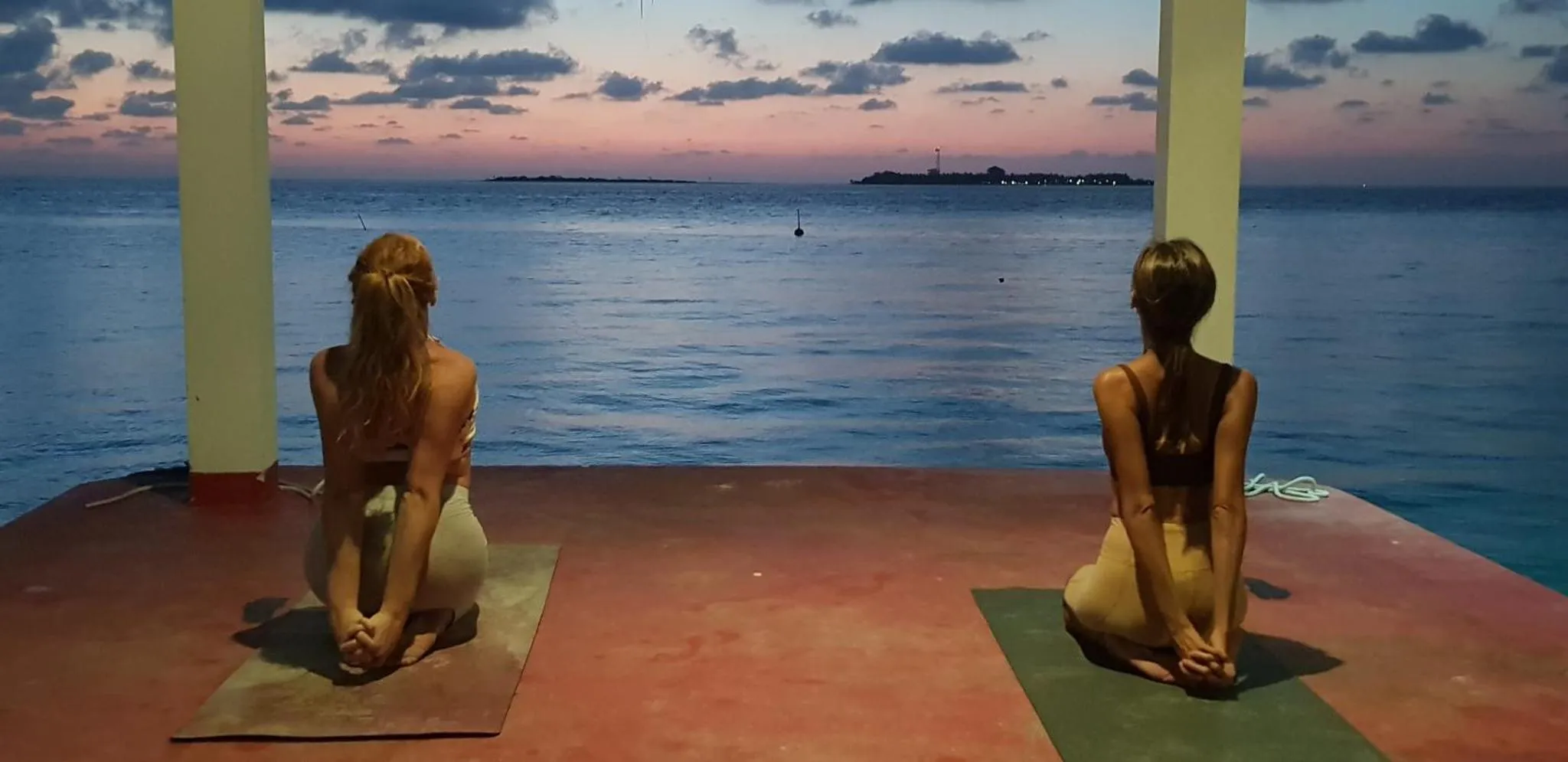 Deux personnes pratiquent le yoga sur une terrasse en bois face à la mer au coucher du soleil.