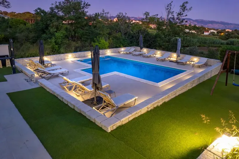 Piscine rectangulaire illuminée avec transats et un espace vert.