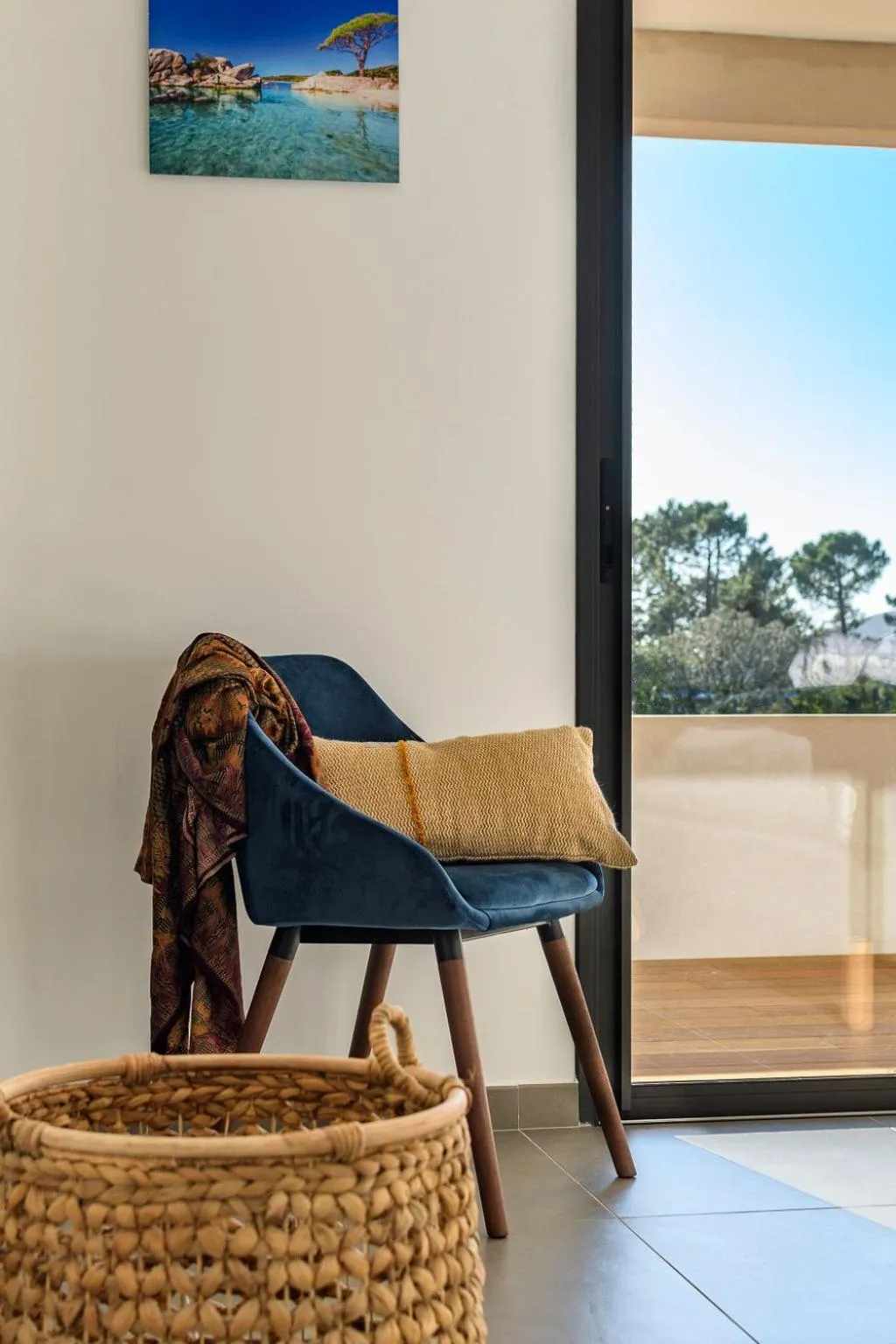 Un fauteuil bleu et un coussin jaune reposent près d'une fenêtre ouverte sur un paysage.