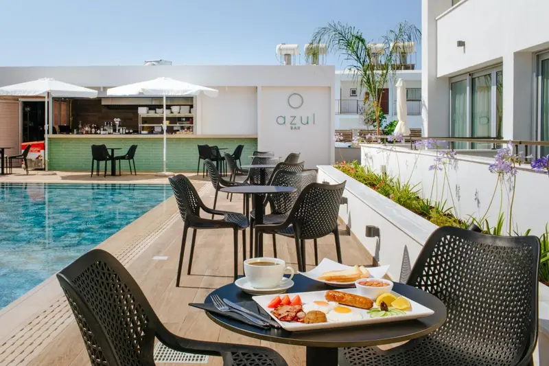 Une table avec un petit-déjeuner est placée au bord d'une piscine avec des transats et un bar en arrière-plan.