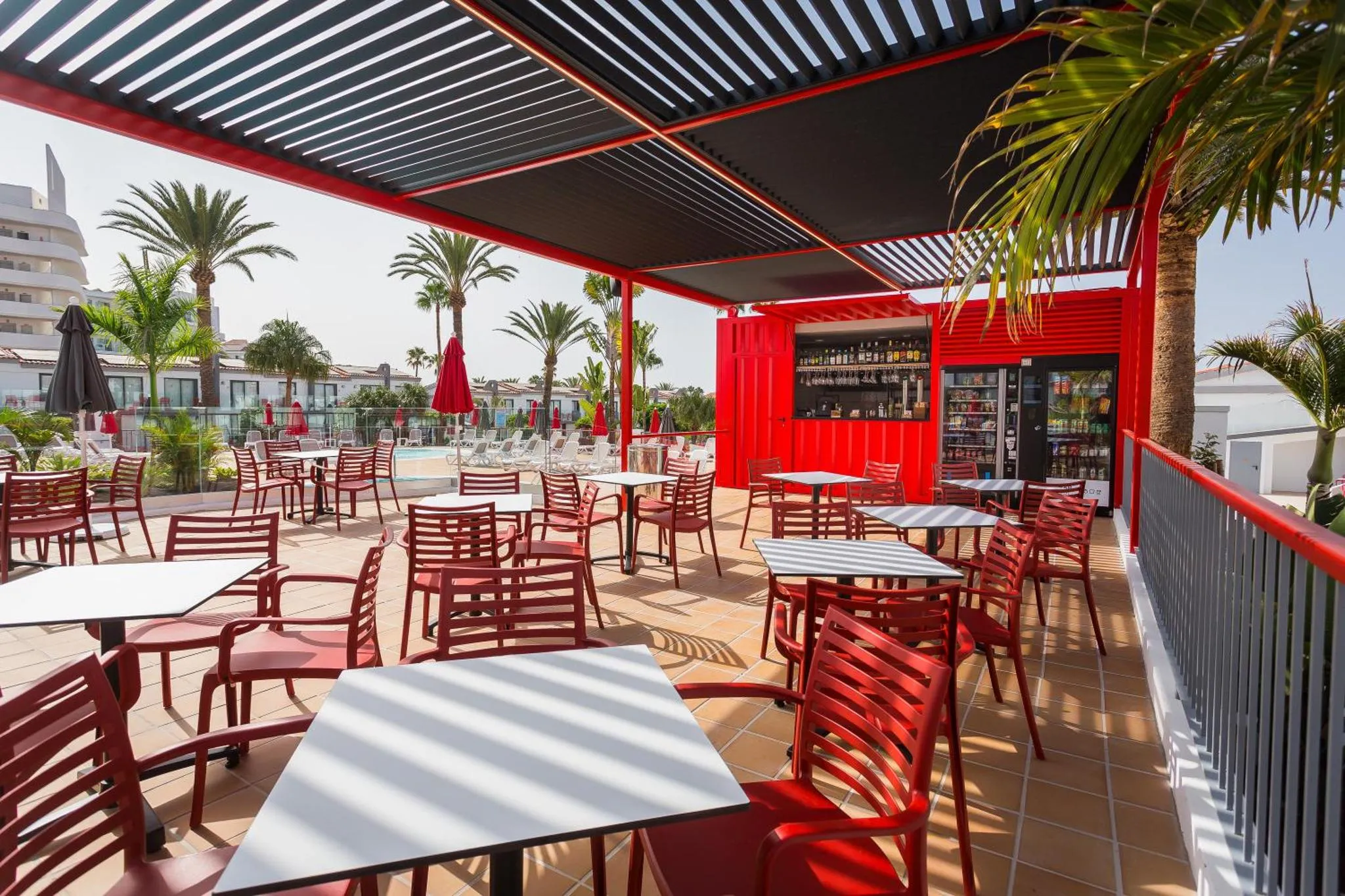 Terrasse ombragée avec tables et chaises rouges, donnant sur une piscine et des palmiers.
