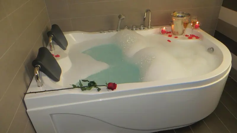 Une baignoire d'angle remplie d'eau et de mousse, décorée de pétales de rose et d'une rose rouge.