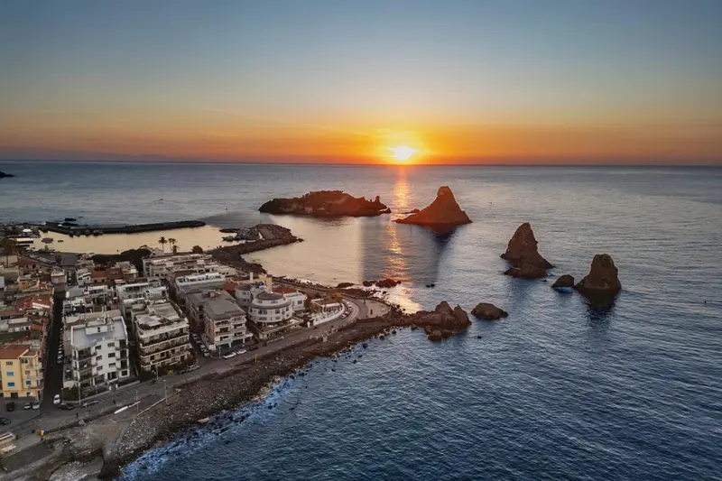Vue aérienne d'une côte rocheuse au coucher du soleil, avec des bâtiments et des formations rocheuses dans la mer.