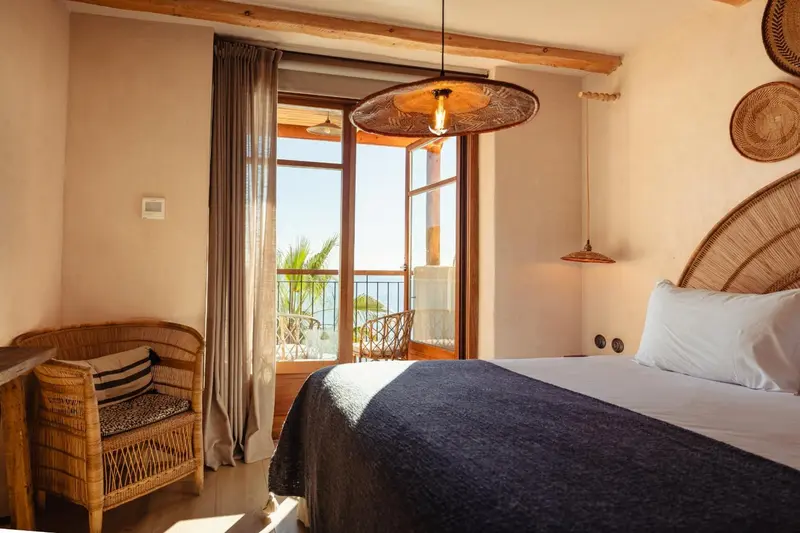 Une chambre lumineuse avec un lit et un fauteuil en osier donnant sur une terrasse.