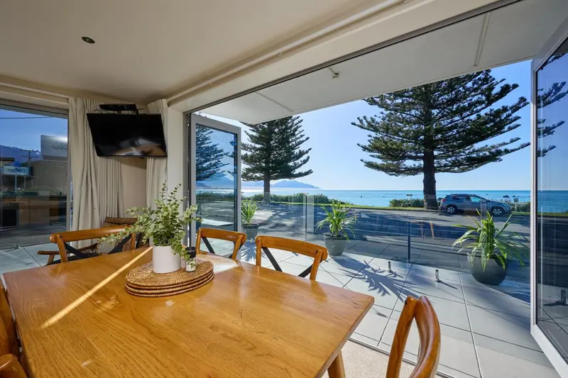 Une table à manger en bois est mise en scène devant une baie vitrée ouverte sur un paysage marin.