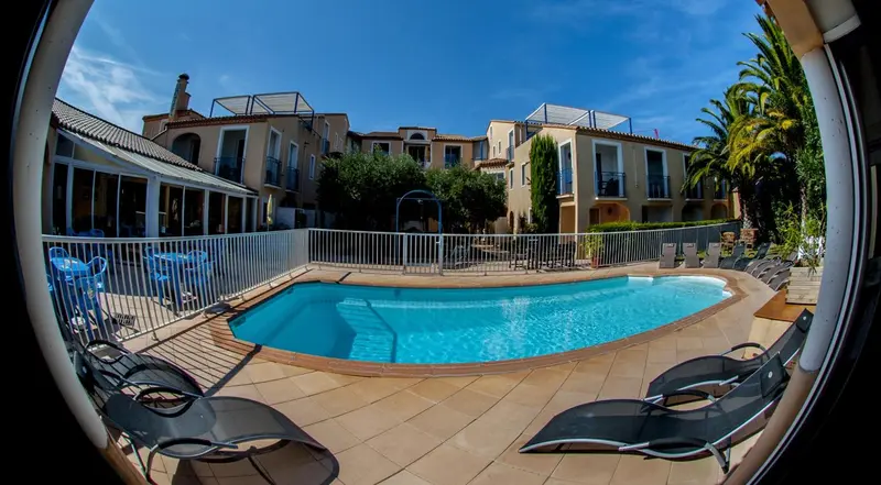 Une piscine turquoise au premier plan, entourée de chaises longues et d'une terrasse carrelée, avec un bâtiment aux façades claires en arrière-plan.