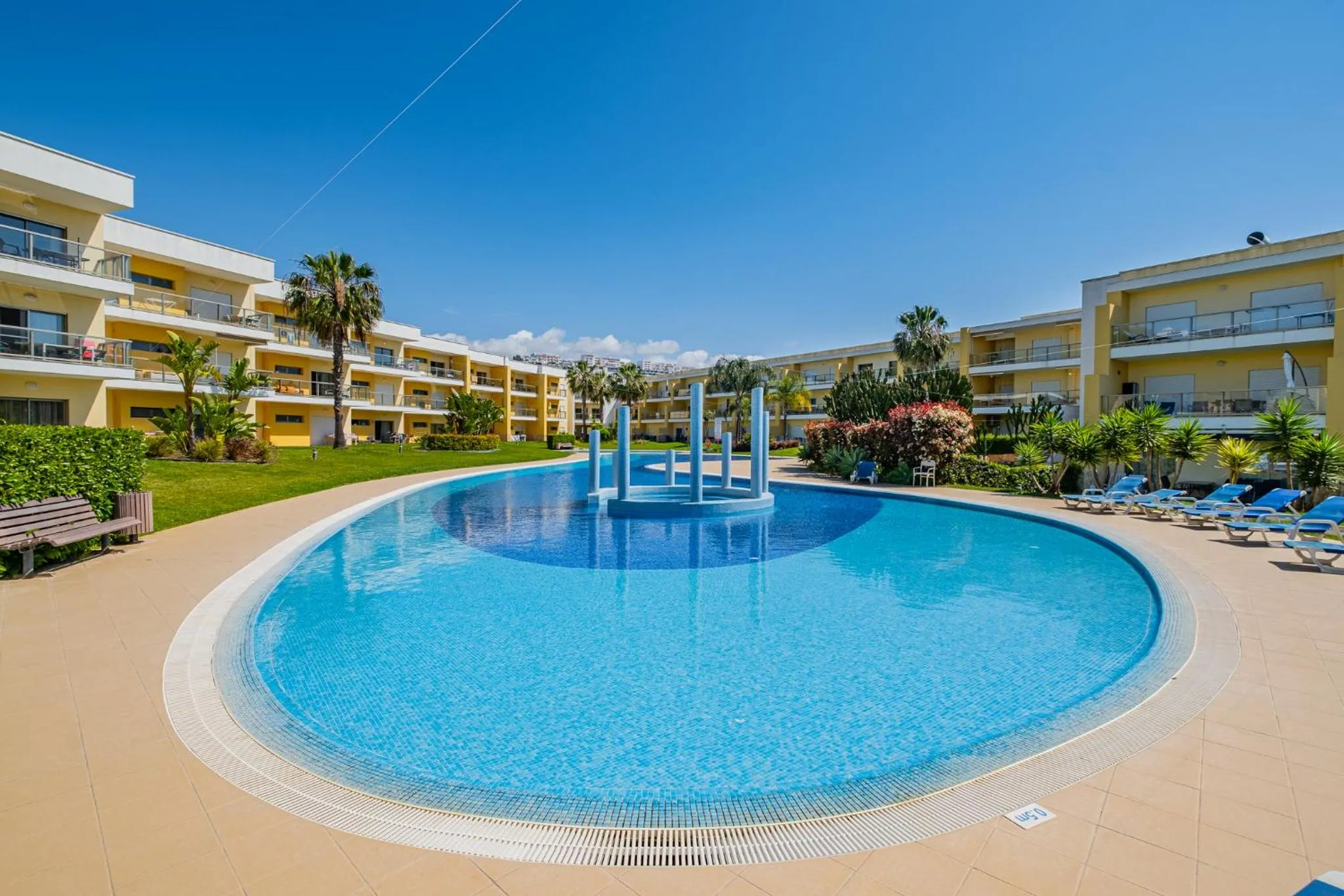 Piscine rectangulaire entourée d'une terrasse ensoleillée avec transats et bâtiments jaunes.