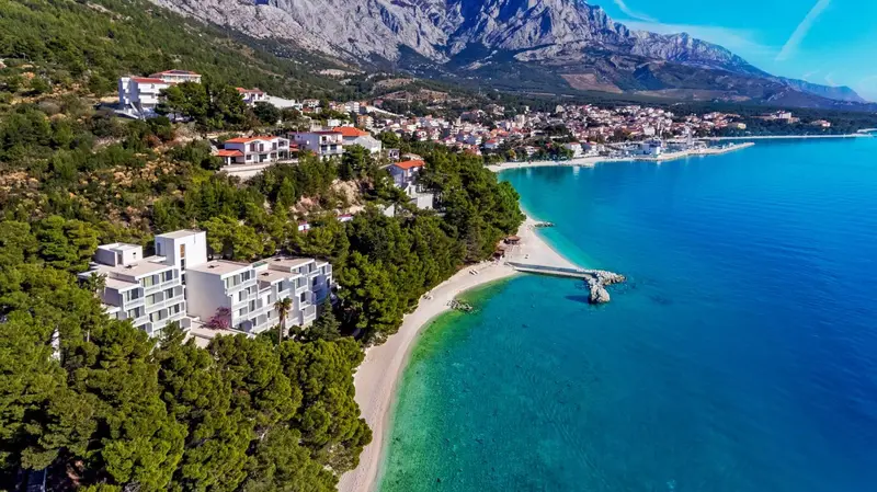 Vue aérienne d'un complexe hôtelier blanc surplombant une plage de galets et une mer turquoise.