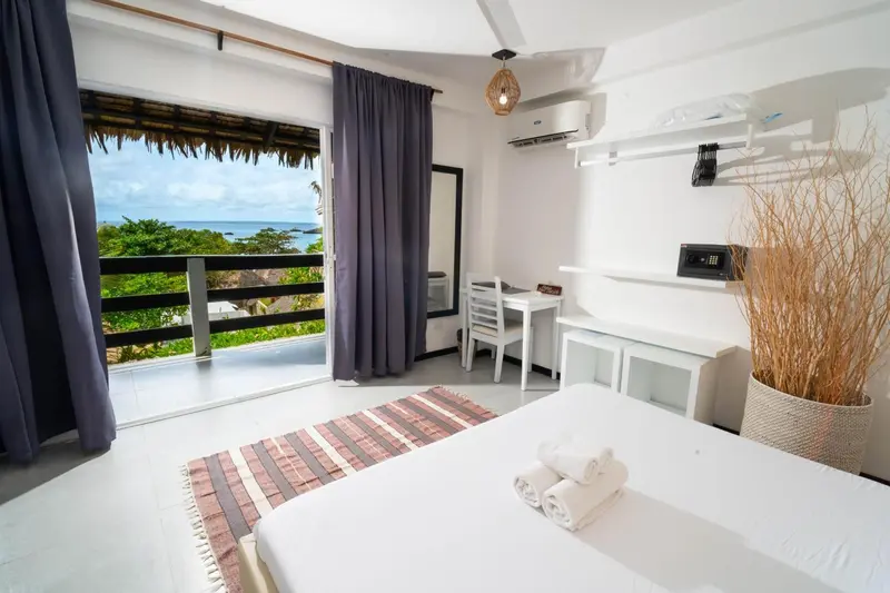 Chambre lumineuse avec un lit blanc et une vue sur la mer depuis une terrasse.