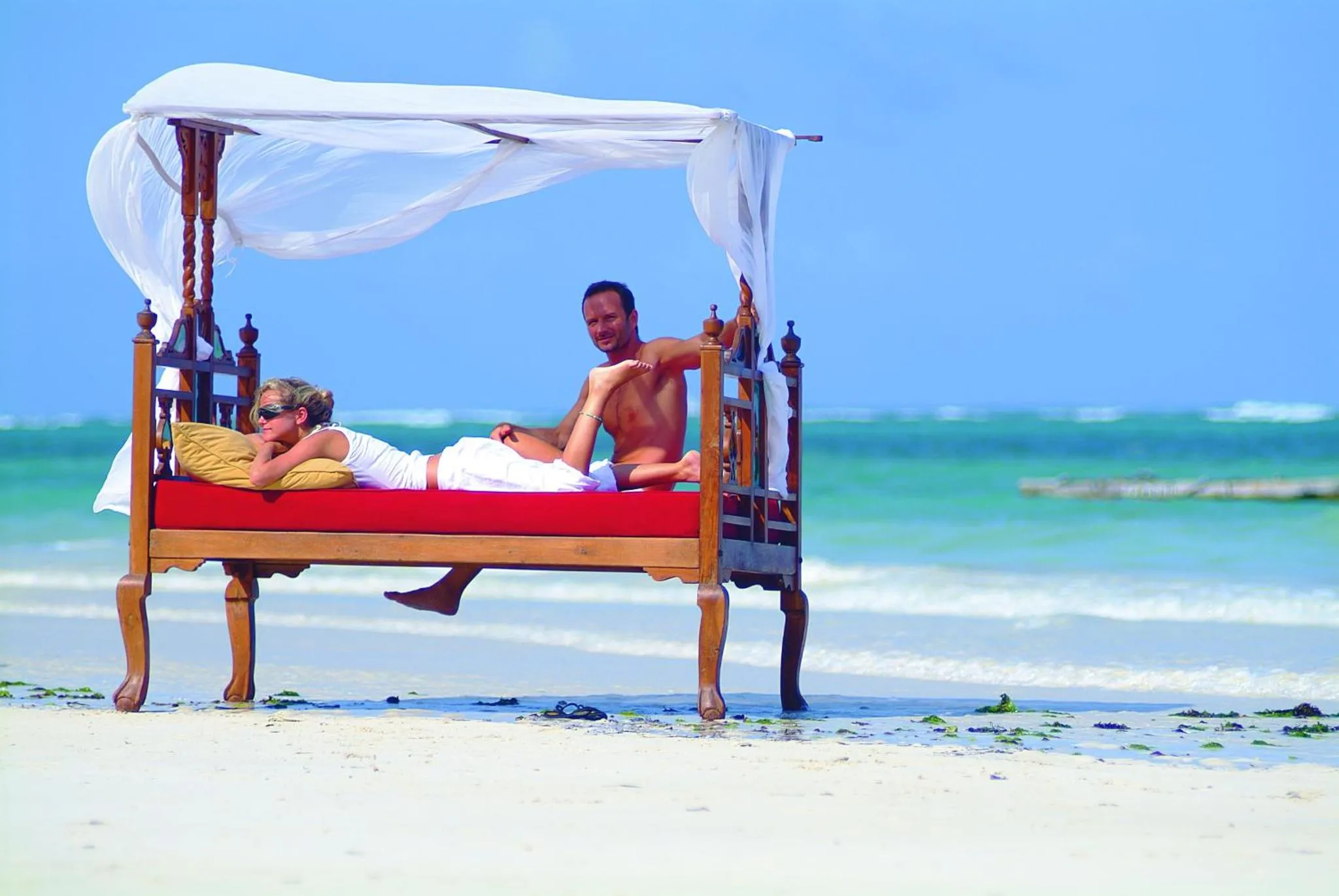 Un couple se repose sur un lit installé sur une plage de sable blanc.