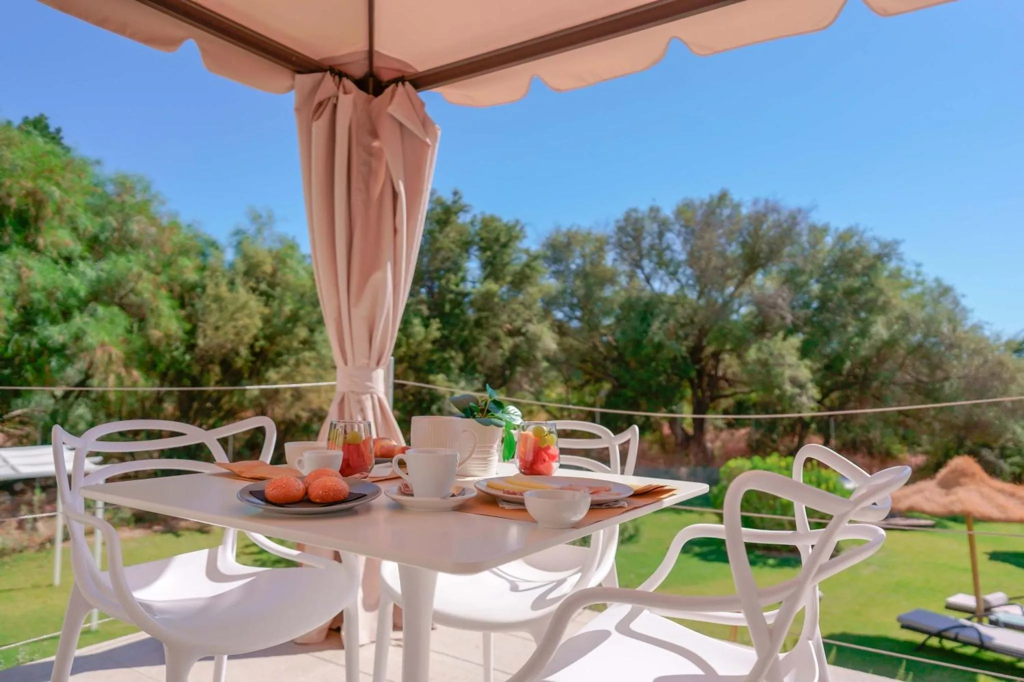 Une table blanche est dressée pour le petit-déjeuner sur une terrasse ombragée.