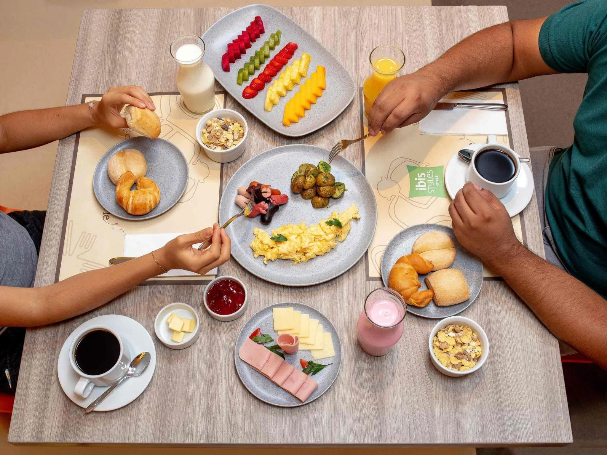 Une table est dressée avec un petit-déjeuner composé de fruits, de yaourts, de viennoiseries et de boissons.