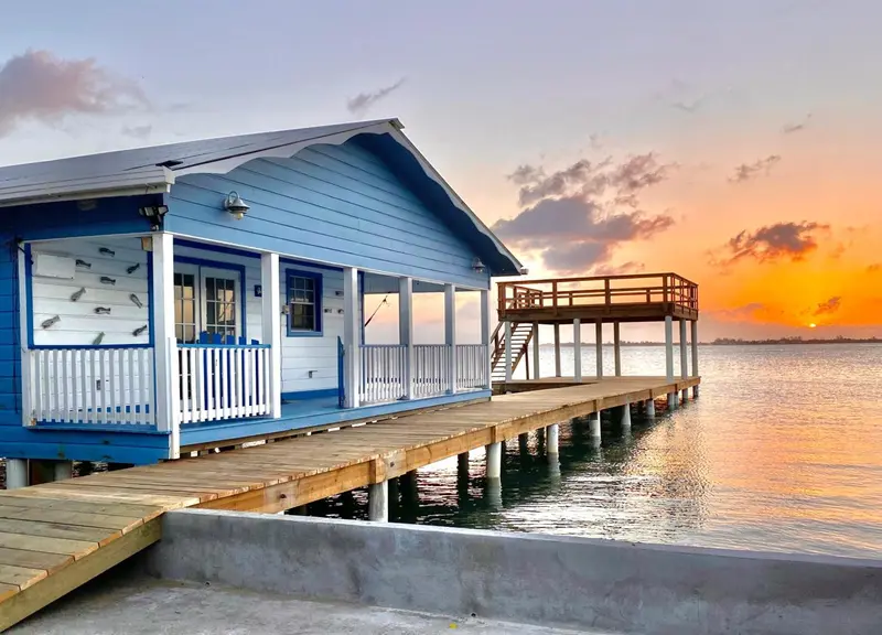 Bâtiment bleu clair sur pilotis avec une terrasse en bois au coucher du soleil.